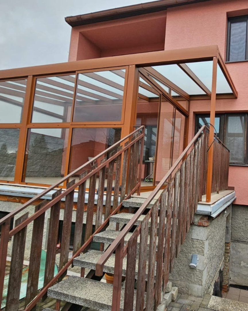 A brown, glass-enclosed aluminum pergola installed on the terrace of a house with a staircase