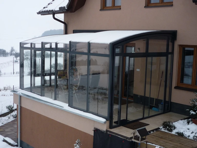 A modern sliding-door sunroom with glass walls, covered in snow, on the patio of a single-family home in winter.