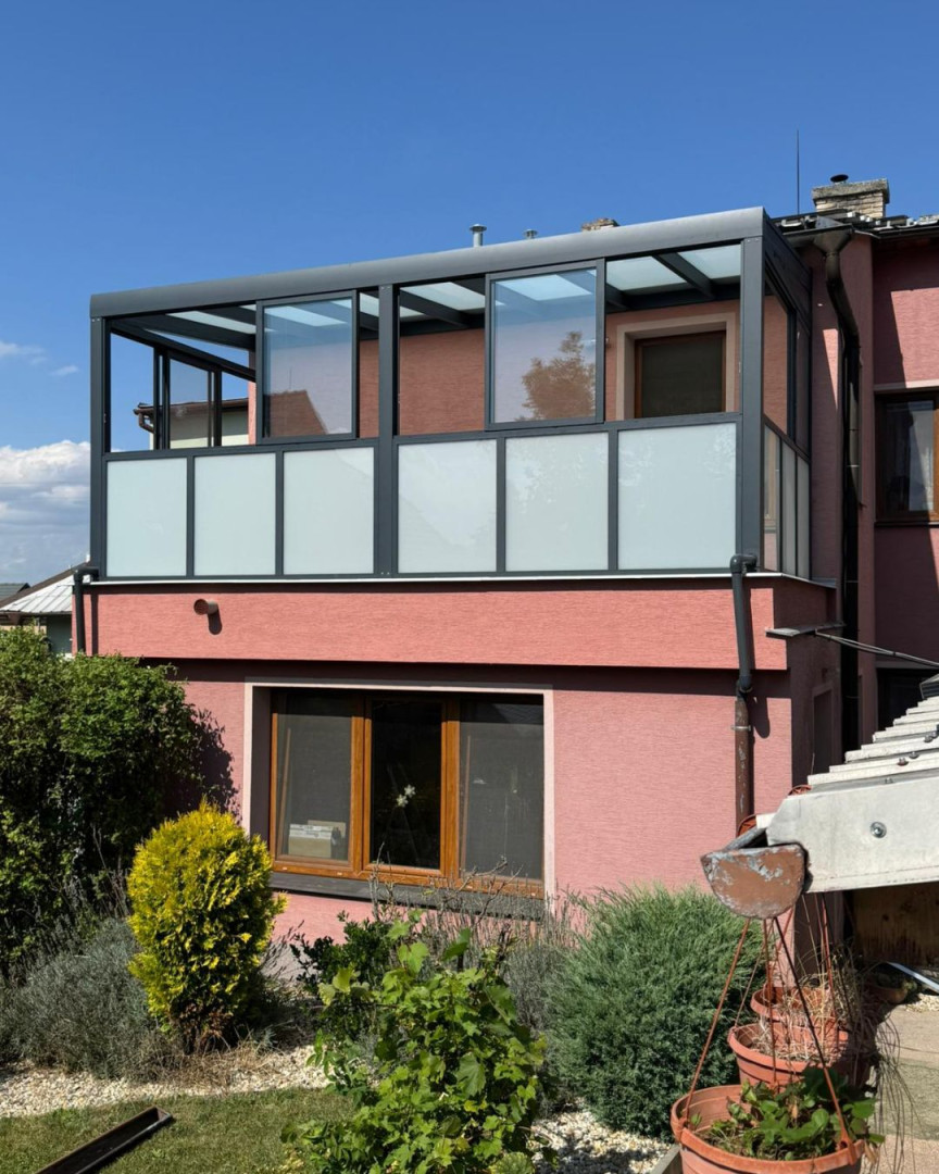 A modern glass pergola with a dark gray aluminum frame and frosted glass on the balcony of a single-family home.