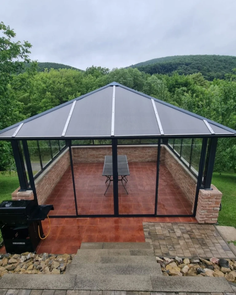 A modern glass gazebo with a dark frame and a brick foundation, set in a natural setting