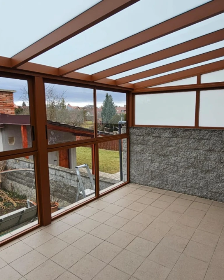 A glass-enclosed aluminum pergola with a brown frame and a view of the garden