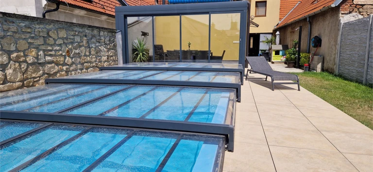 A modern, low-profile telescopic pool enclosure with an anthracite aluminum frame on the terrace next to the house.
