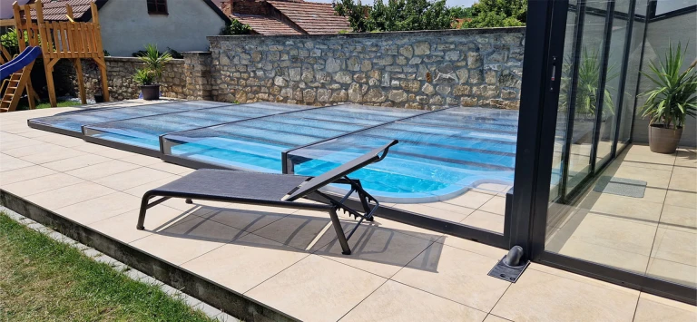 A modern, low-profile pool enclosure with a black aluminum frame on a patio with a lounge chair