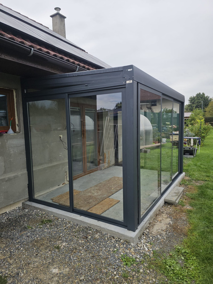 A modern, glass-enclosed conservatory addition with a dark gray frame attached to a single-family home