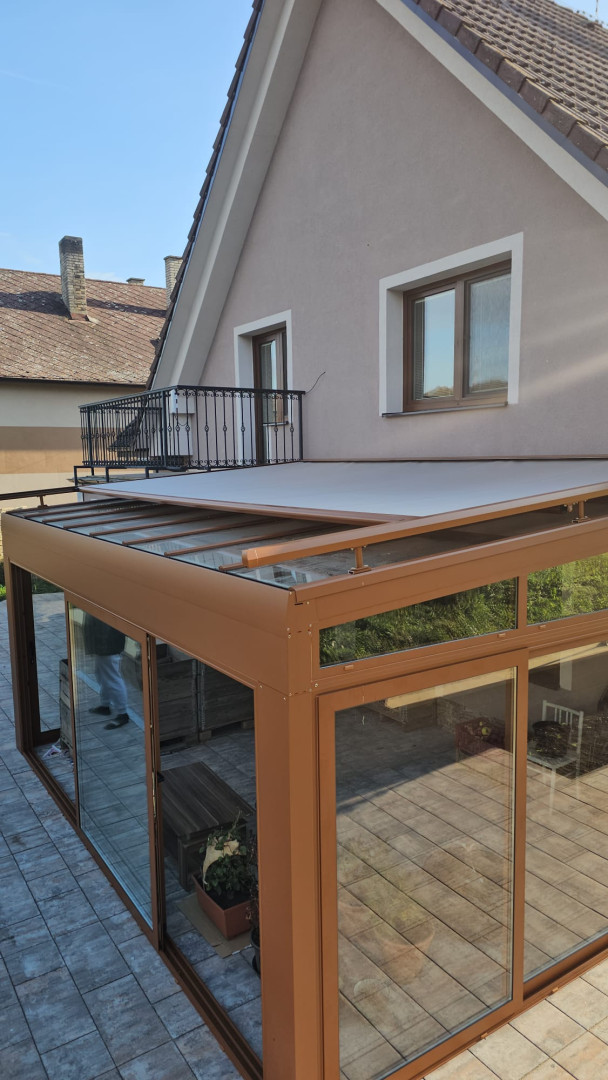 A glass-enclosed sunroom with a brown frame and a retractable awning on the terrace of a single-family home
