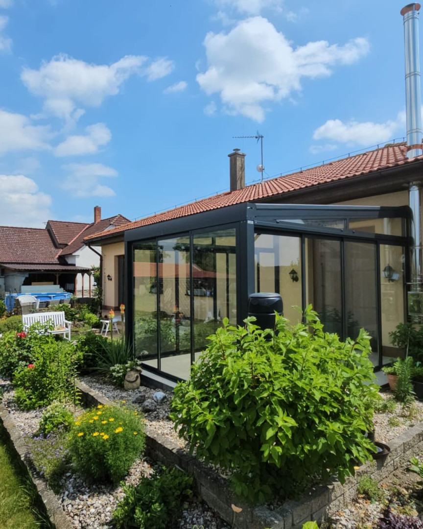 A modern, glass-enclosed sunroom with an anthracite-colored frame attached to a single-family home with a well-maintained garden