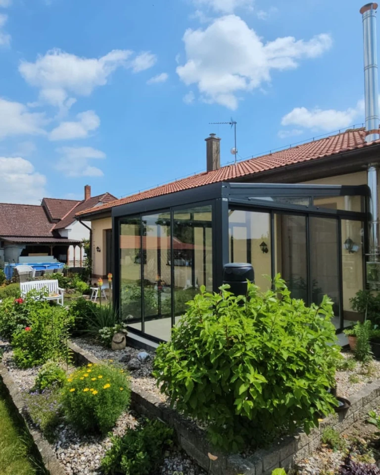 A modern, glass-enclosed sunroom with an anthracite-colored frame attached to a single-family home with a well-maintained garden