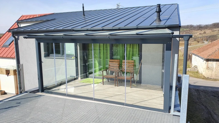 A modern glass-enclosed sunroom with an anthracite-colored frame and sliding glass doors on the house's terrace.